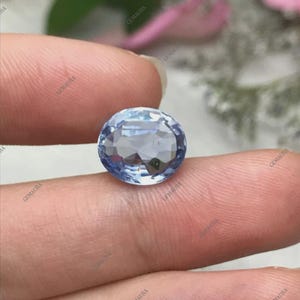 May include: A faceted, oval-cut, light blue gemstone. The gem is held between two fingers, showcasing its clarity and brilliance. The stone's facets reflect light, creating a sparkling effect. The background is blurred, focusing attention on the gemstone.