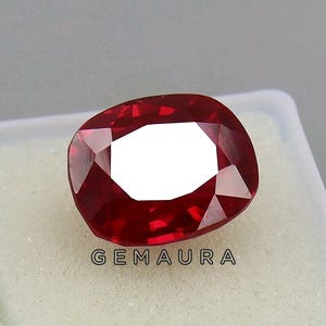 May include: A faceted, cushion-cut ruby gemstone in a deep red colour. The gem is set against a white background, with the text "GEMAURA" visible at the bottom. The gemstone's facets reflect light, creating a sparkling effect.