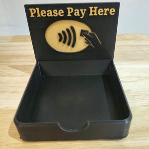 May include: A black payment tray with a raised back displaying the text "Please Pay Here" in gold. Below the text is a gold oval with a contactless payment symbol and a hand holding a card. The tray is empty.