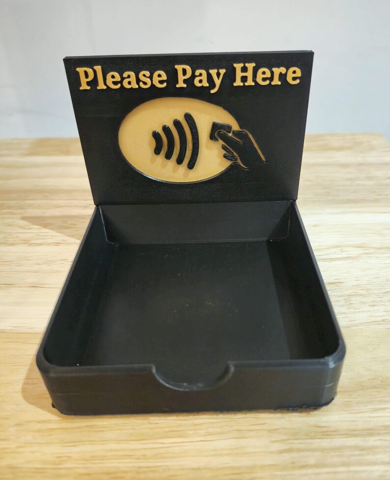 May include: A black payment tray with a raised back displaying the text "Please Pay Here" in gold. Below the text is a gold oval with a contactless payment symbol and a hand holding a card. The tray is empty.