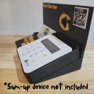 May include: A black display stand holding a white SumUp device. The stand features a slot for business cards and a QR code. The text "Please Take One" is displayed on the stand.