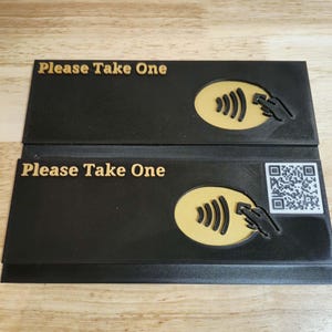 May include: Two black rectangular signs with the text "Please Take One" in gold. Each sign features a gold contactless payment symbol and a hand icon. One sign also has a QR code.