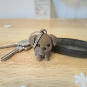 May include: A brown dog keyring with a silver ring. The dog is a small, 3D-printed figurine with black eyes. Keys and a car remote are in the background. The keyring is on a wooden surface. A charming accessory.