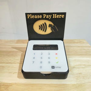 May include: A black and white SumUp card reader with a display and number pad. The reader has a hinged top with the words "Please Pay Here" and a contactless payment symbol. The reader is on a wooden surface.