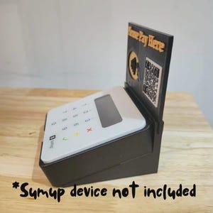 May include: Black payment terminal stand with a white keypad and a display screen. The stand has a sign that says "Please Pay Here" with a QR code. The text "Sumup device not included" is at the bottom.