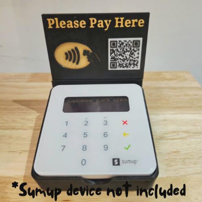 May include: A white and black payment terminal with a numeric keypad and a display screen. The terminal has a black stand with the text "Please Pay Here" and a QR code. The image also includes the text "Sumup device not included".