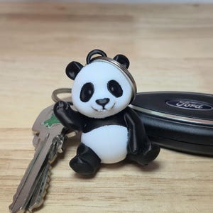 May include: A black and white panda bear keychain with a silver key ring. The panda is sitting and waving. A car key and a Ford key fob are in the background.