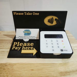 May include: A black and gold point-of-sale display with the text "Please Take One" and "Please Pay here". A white SumUp card reader is in the display. A business card is also in the display.