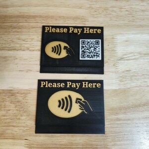 May include: Two black rectangular signs with the text "Please Pay Here" in gold. One sign has a QR code and a contactless payment symbol. The other sign has the contactless payment symbol. Both signs are on a wooden surface.