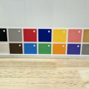 May include: A white plastic holder displaying a variety of small, square colour samples. The samples include black, grey, red, blue, yellow, pink, and gold, along with other shades. Each square has a small hole. The holder sits on a wooden surface.