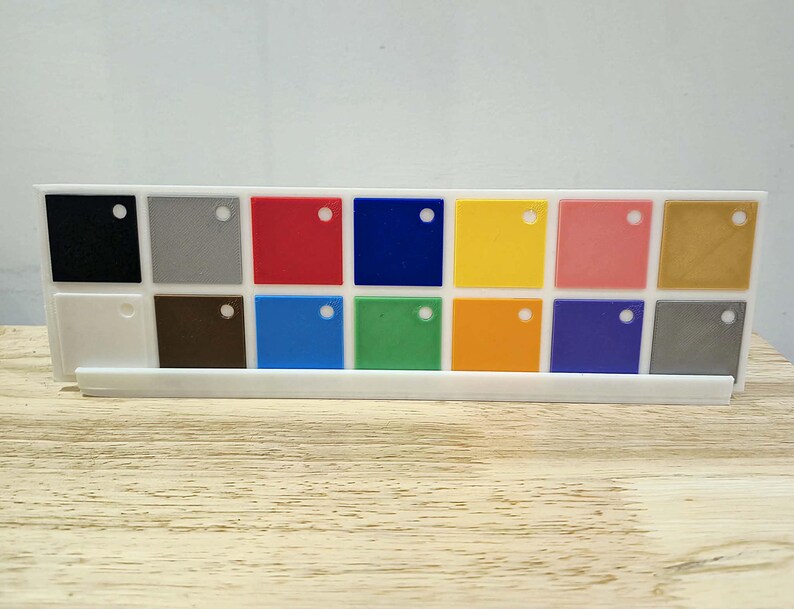 May include: A white plastic holder displaying a variety of small, square colour samples. The samples include black, grey, red, blue, yellow, pink, and gold, along with other shades. Each square has a small hole. The holder sits on a wooden surface.