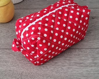May include: Red quilted cosmetic bag with white polka dots and a white zipper. The bag is rectangular and has a small fabric loop on one end. The bag is sitting on a wooden surface.