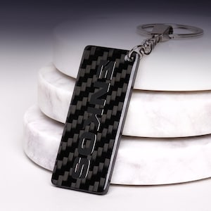 Peut inclure: Un porte-clés rectangulaire arborant un motif en fibre de carbone noir et gris avec le mot ENKOS embossé verticalement dans une police sombre et épaisse. Il est fixé à un anneau brisé en métal argenté par une courte chaîne torsadée.