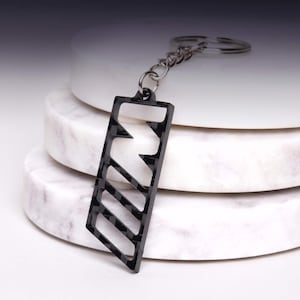 Op de afbeelding: Een zwarte sleutelhanger met het gestileerde M-logo in een koolstofvezelpatroon. De sleutelhanger is met een kort kettinkje bevestigd aan een zilverkleurige metalen ring. Het ontwerp bestaat uit drie schuine balken die de letter M vormen.