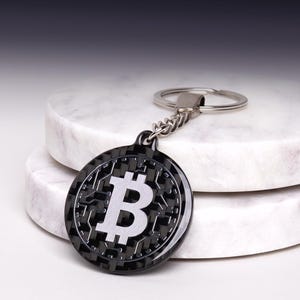 May include: A circular keyring featuring a black carbon fibre-style background with a prominent silver Bitcoin symbol in the centre. The design includes a metallic circuit board pattern surrounding the logo, attached to a silver metal split ring.