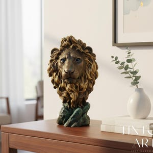 May include: A bronze lion head sculpture with a detailed mane and blue eyes. The sculpture sits on a green, textured base. The lion is displayed on a wooden table, with a white vase and books in the background.