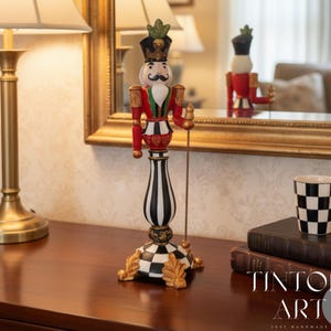 Puede incluir: Figura decorativa de cascanueces con base a rayas blancas y negras, chaqueta roja y detalles dorados. La figura está sobre una mesa junto a una lámpara y una taza a cuadros. La figura se refleja en un espejo con marco dorado.