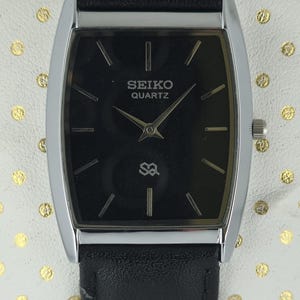 Könnte beinhalten: Eine rechteckige Seiko Quartz-Uhr mit schwarzem Zifferblatt und silberfarbenem Gehäuse. Die Uhr hat ein schwarzes Lederarmband und das Seiko-Logo. Sie hat silberne Zeiger und Markierungen. Die Uhr wird auf einer weißen Oberfläche mit goldenen Punkten präsentiert.