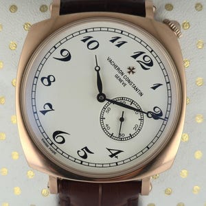 Puede incluir: Un reloj de pulsera de oro rosa con esfera crema y números negros. El reloj tiene un pequeño dial de segundos y una correa de cuero marrón. La marca "VACHERON CONSTANTIN GENEVE" está impresa en la esfera. Fabricado en Suiza.