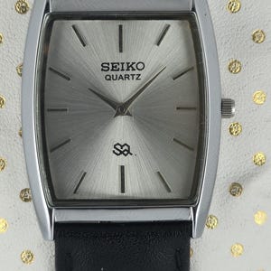 Może przedstawiać: Prostokątny zegarek Seiko Quartz ze srebrną tarczą i czarnym skórzanym paskiem. Tarcza zegarka ma srebrne wykończenie z czarnymi liniami wskazującymi godziny. Zegarek leży na białej powierzchni ze złotymi kropkami.