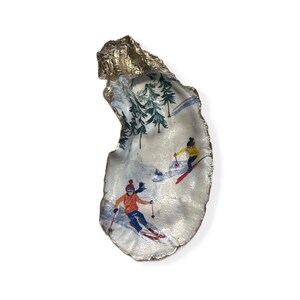 May include: A gold oyster shell with a painted scene of two skiers on a snowy mountain slope with pine trees.