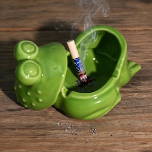 Puede incluir: Un cenicero de cerámica verde con forma de rana con un cigarrillo encendido. El cenicero está sobre una superficie de madera y sale humo del cigarrillo. El cenicero tiene un acabado brillante.