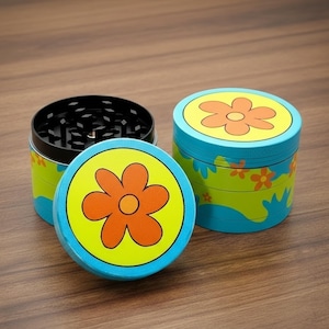 May include: A set of three-piece herb grinders with a flower design. The grinders have a blue, yellow, and green color scheme. The top of the grinder has a flower design with an orange flower on a yellow background. The grinder is made of metal.
