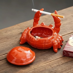May include: A glossy, orange crab-shaped ashtray with a matching lid. The ashtray holds a lit cigarette, and another cigarette rests in the crab's claws. A pack of cigarettes sits nearby on a wooden surface.