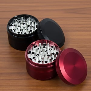 May include: Two herb grinders, one black and one red, sit on a wooden surface. The red grinder has its lid off, revealing the metal grinding teeth. The black grinder has its lid on. The lids are circular and the grinders are cylindrical.