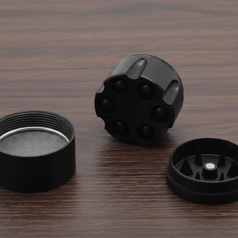 May include: A black, disassembled herb grinder with a revolver-style top. The grinder is made of metal and has a mesh screen and a bottom piece with teeth. The pieces are arranged on a dark wood surface.