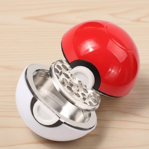 Peut inclure: Un broyeur fantaisie rouge et blanc, conçu pour ressembler à une balle d'un personnage de dessin animé populaire. Le broyeur est ouvert, révélant un composant de broyage en métal argenté. L'article est sur une surface en bois clair.