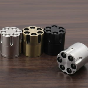 May include: Four metal grinders in silver, gold, and black, arranged on a wooden surface. Each grinder has a cylindrical body and a top with multiple circular openings, resembling a revolver cylinder. One grinder is positioned on its side.