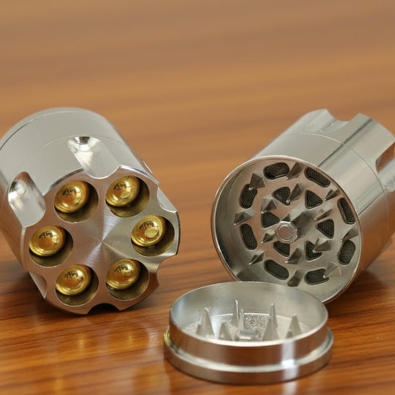May include: A silver-colored, three-piece herb grinder with a revolver cylinder design. The grinder is disassembled, revealing the grinding teeth and the chamber for ground herbs. The cylinder features six gold-colored bullet-shaped chambers.