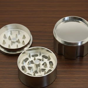 May include: A three-piece, silver-colored herb grinder. The grinder is disassembled, showing the top, bottom, and middle sections. The bottom and middle sections have sharp, triangular teeth for grinding. The top section is a lid.