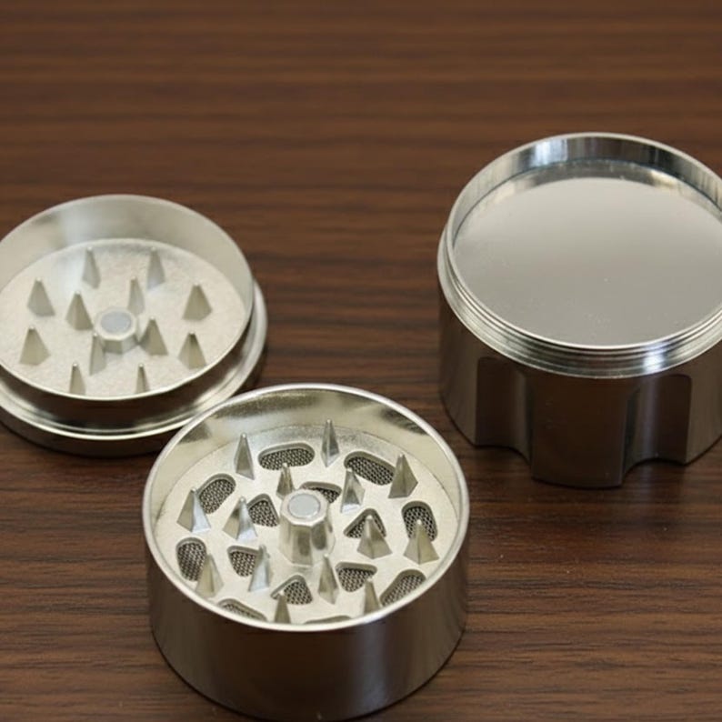 May include: A three-piece, silver-colored herb grinder. The grinder is disassembled, showing the top, bottom, and middle sections. The bottom and middle sections have sharp, triangular teeth for grinding. The top section is a lid.