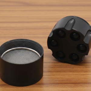 May include: A black, disassembled grinder with a mesh screen and a revolver-cylinder-shaped top. The grinder is made of metal and sits on a wooden surface. The top has six circular indentations.