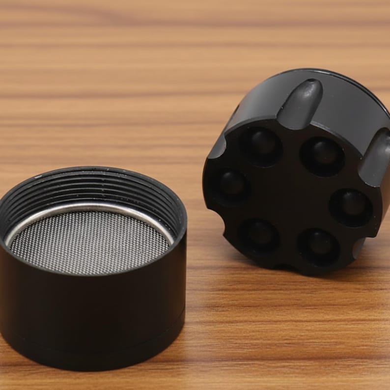 May include: A black, disassembled grinder with a mesh screen and a revolver-cylinder-shaped top. The grinder is made of metal and sits on a wooden surface. The top has six circular indentations.