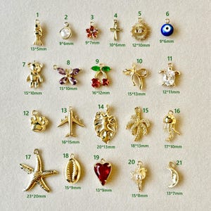 May include: A selection of gold-coloured charms in diverse shapes, such as a fish, pearl, flower, cross, eye, bear, butterfly, cherries, bow, turtle, paw print, airplane, leaf, palm tree, jellyfish, starfish, shell, heart, and moon.