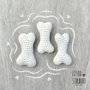 Op de afbeelding: Drie witte, gehaakte hondenbotten op een grijze houten ondergrond. De botten hebben een klassieke botvorm, met een getextureerd, handgemaakt uiterlijk. De afbeelding bevat de tekst "Lusha Studio Shop".