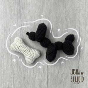 May include: A crocheted black dog balloon animal and a white crocheted bone are displayed on a light gray wooden surface. The dog balloon animal is made of black yarn and has a rounded shape. The bone is white and has a classic bone shape.