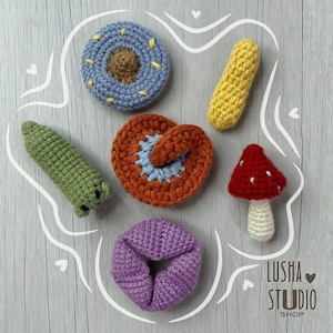 Könnte beinhalten: Eine Sammlung handgefertigter gehäkelter Lebensmittel, darunter ein blauer Donut, ein grüner Maiskolben, ein gelber Hotdog, eine orangefarbene Brezel, ein roter Pilz und ein lila Scrunchie. Die Artikel sind auf einer hellgrauen Holzoberfläche angeordnet.