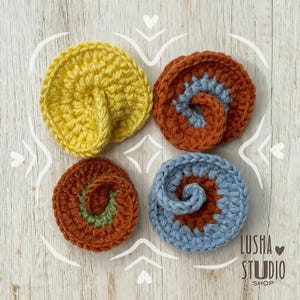 Pode incluir: Quatro bases de copo de crochê feitas à mão em tons de amarelo, ferrugem e azul. Cada base de copo apresenta um design em espiral. As bases de copo são dispostas sobre uma superfície de madeira clara com detalhes brancos decorativos. O nome da loja "Lusha Studio Shop" é visível.