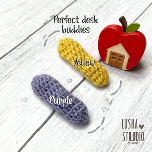 May include: A wooden apple-shaped desk accessory with a house cutout, a yellow crocheted item, and a purple crocheted item. The text "Perfect desk buddies" and "Yellow" and "Purple" are visible.
