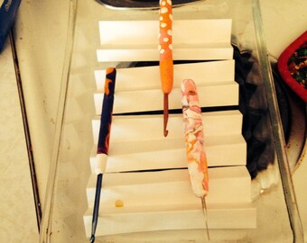 May include: Three crochet hooks in a glass baking dish. The hooks are made of polymer clay and are decorated with white dots, pink and white swirls, and black and white stripes.