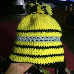 May include: A yellow and black striped crocheted hat with a grey and black pom-pom on top. The hat has a black crocheted brim and ear flaps.
