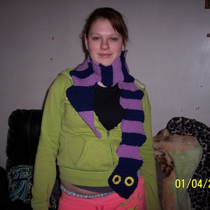 May include: A purple and blue striped crocheted scarf shaped like a snake with yellow eyes. The scarf is worn by a person with brown hair and a green sweater.