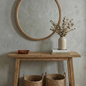 Könnte beinhalten: Ein Holztisch mit einem runden Spiegel, einer Vase mit getrockneten Pflanzen, Büchern und zwei geflochtenen Körben. Der Tisch hat eine natürliche Holzoberfläche. Der Spiegel hat einen Holzrahmen. Die Körbe sind aus gewebtem Material.