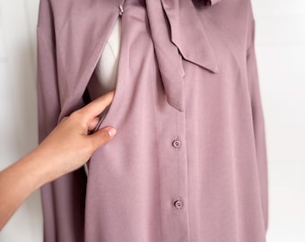 Puede incluir: Una blusa color malva con un gran lazo en el cuello y mangas largas. La camisa abotonada se exhibe en un maniquí. Una mano sostiene el lateral de la blusa.