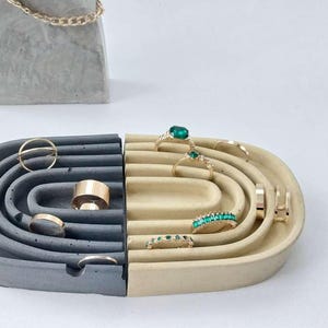 Könnte beinhalten: Ein zweifarbiges Schmuck-Organizer aus Beton in Grau und Beige. Der Organizer hat gebogene Rillen für Ringe und einen separaten Ständer für Halsketten. Goldringe und eine goldene Kette werden präsentiert.