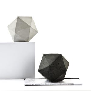 May include: Two geometric concrete sculptures. One is light gray and rests on a white block. The other is dark gray and sits on a rectangular, speckled surface. Both have a multifaceted, diamond-like shape.